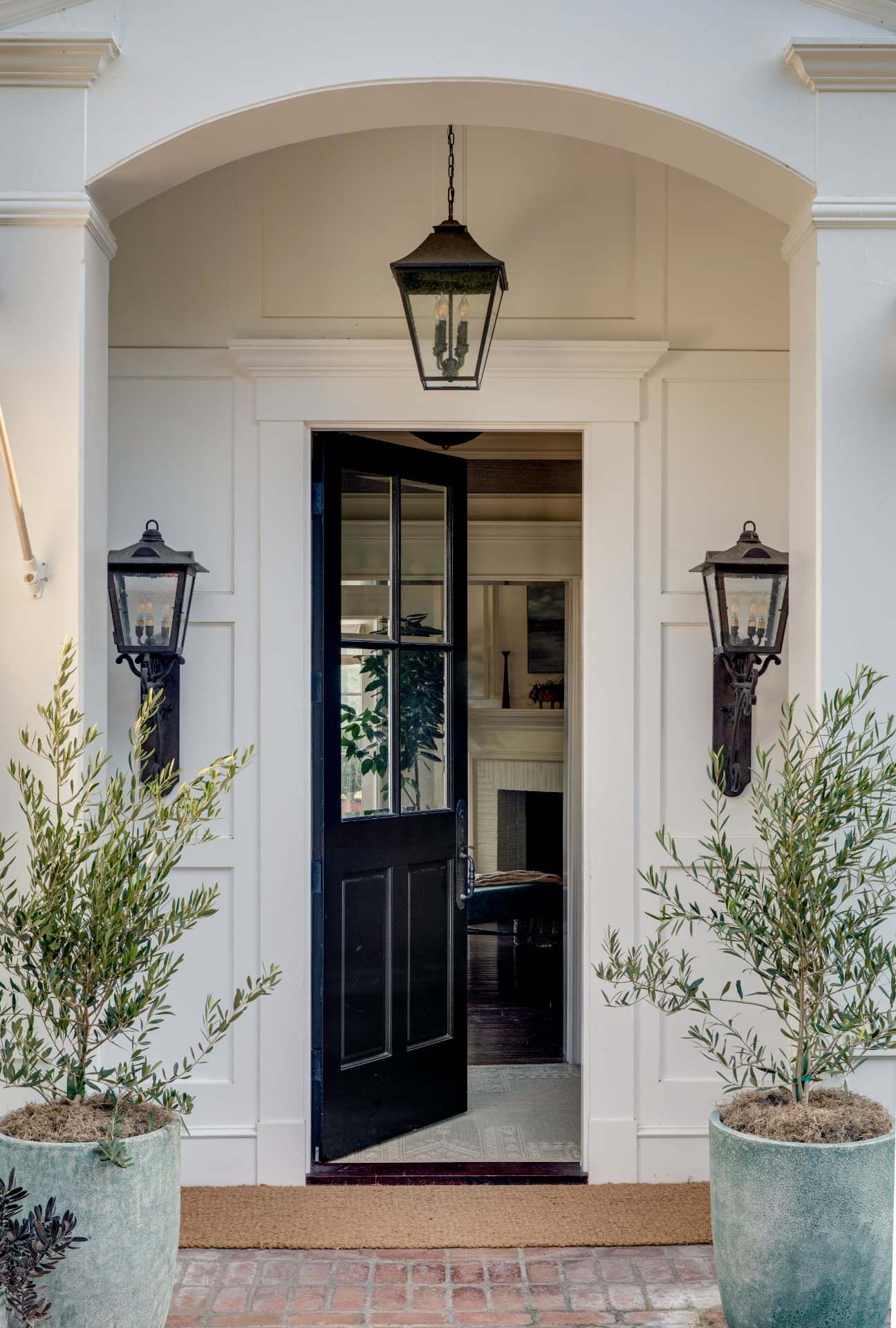 Arched entry porch with lanterns, black door, and potted olive trees